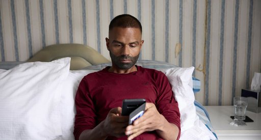 A man sitting on a bed, looking at a mobile phone.