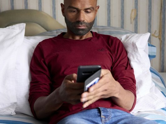 A man sitting on a bed, looking at his phone.