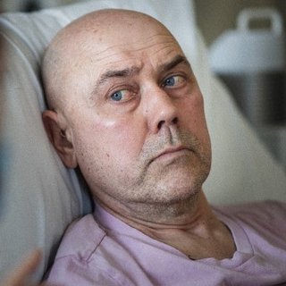 Image of a white man in bed in hospital. He is looking at a nurse.