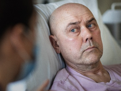 Image of a white man in bed in hospital. He is looking at a nurse.