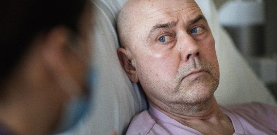 Image of a white man in bed in hospital. He is looking at a nurse.
