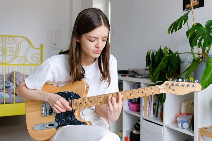 Someone wearing white t-shirt looking downwards playing brown electric guitar in bedroom.