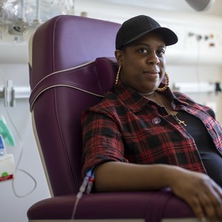 A woman sitting in a hospital receiving intravenous treatment.   She's sitting in a chair, with a thin plastic tube coming out from under her rolled up shirt sleeve. There's an IV stand next to her.