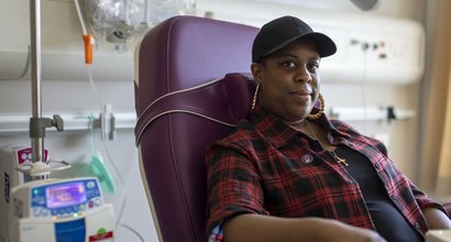 A woman sitting in a hospital receiving intravenous treatment.   She's sitting in a chair, with a thin plastic tube coming out from under her rolled up shirt sleeve. There's an IV stand next to her.
