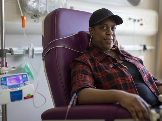 A woman sitting in a hospital receiving intravenous treatment. She's sitting in a chair, with a thin plastic tube coming out from under her rolled up shirt sleeve. There's an IV stand next to her.