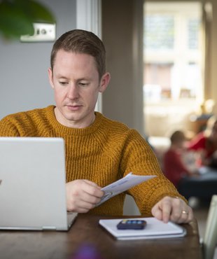 A man - Rob - carries out some admin at a computer, sat at his dining table. A calculate and notepad are at hand.