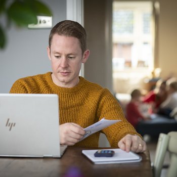 A man - Rob - carries out some admin at a computer, sat at his dining table. A calculate and notepad are at hand.