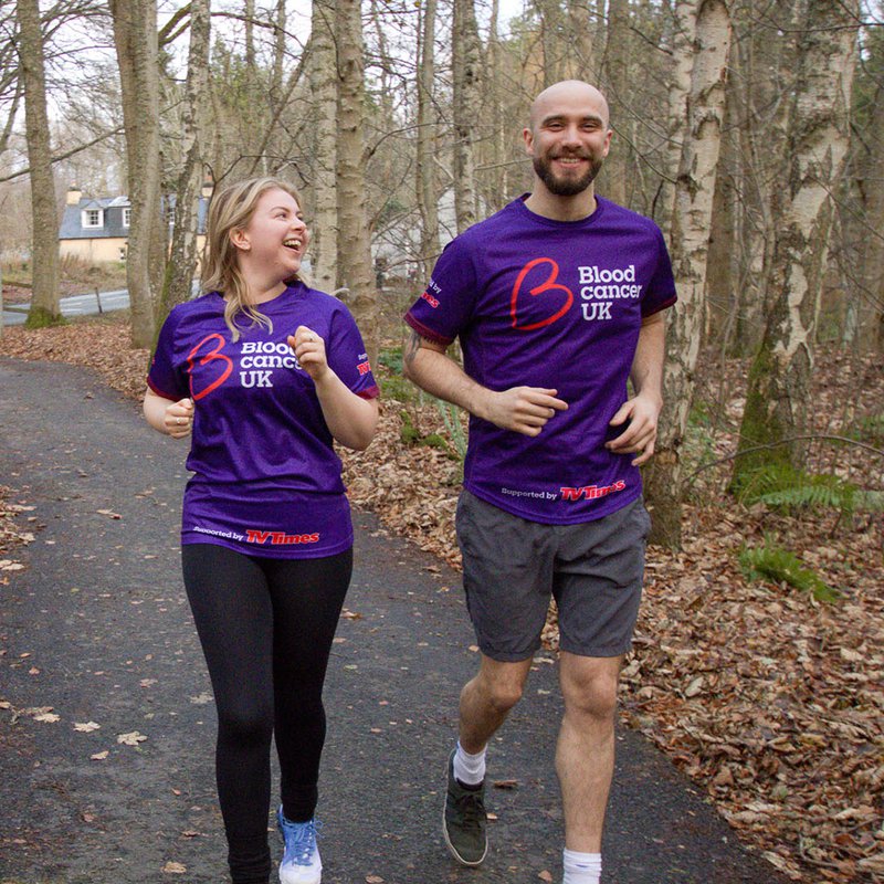 Two people run through the woods wearing purple Blood Cancer UK t-shirts