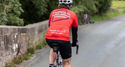 A cyclist heading down a country lane, wearing a Blood Cancer UK T shirt with "Cycling to beat blood cancer' on the back.