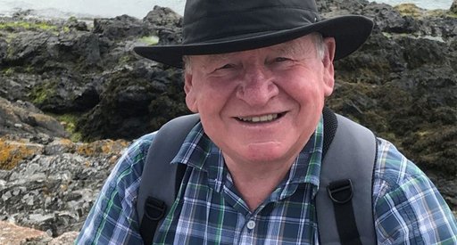 An older man - Gerald - in the countryside. He wears a brimmed hat, check shirt and has a backpack. The image is closely cropped with Gerald looking direct to camera and smiling.