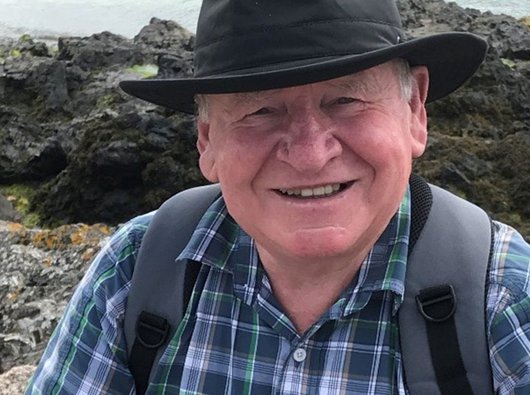 An older man - Gerald - in the countryside. He wears a brimmed hat, check shirt and has a backpack. The image is closely cropped with Gerald looking direct to camera and smiling.