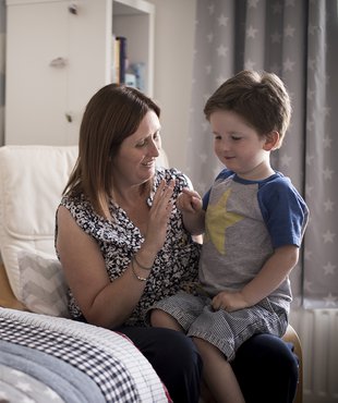 A child of about 3 shares a tender moment with his mother, propped on her knee and looking towards a grouping of cuddly toys.