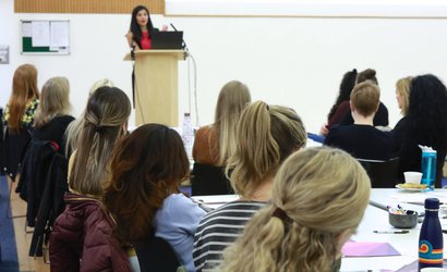 At a meeting, a woman stands and speaks with a laptop while an all-female seated crowd listens.