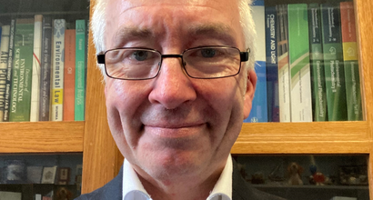 Photo of Peter, smiling and wearing a blazer standing in front of his book shelf.
