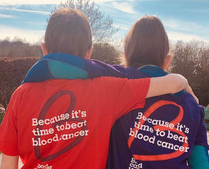 Two supporters hug, wearing Blood Cancer UK T-shirts, with the slogan "Because it's time to beat blood cancer"