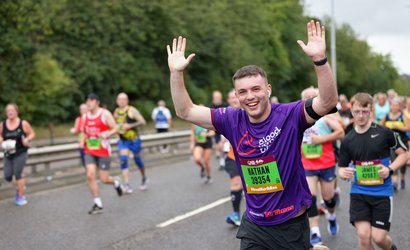 A man cheers and raises his hands in the air as he runs through the city for the Great North Run, wearing a purple Blood Cancer UK running vest