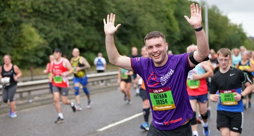 A man cheers and raises his hands in the air as he runs through the city for the Great North Run, wearing a purple Blood Cancer UK running vest