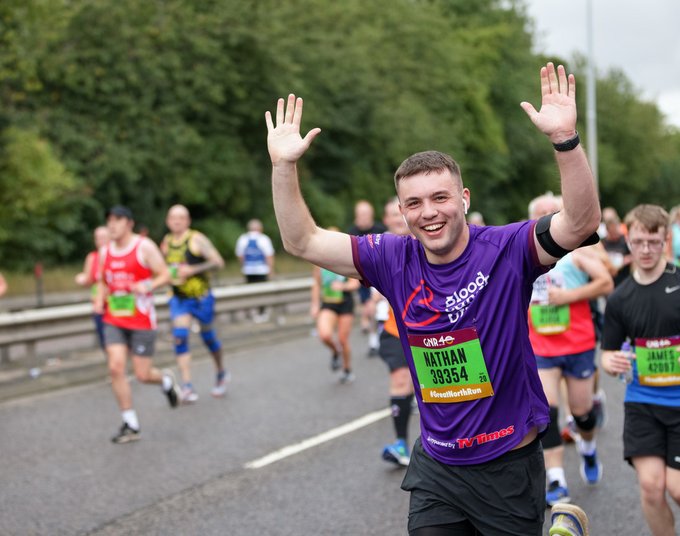 A man cheers and raises his hands in the air as he runs through the city for the Great North Run, wearing a purple Blood Cancer UK running vest
