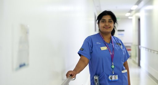 A close up of a nurse, smiling in her blue uniform, standing in the corridor of a hospital.