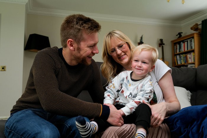Emma sits with her husband and her baby son on the sofa, they're smiling and look happy together