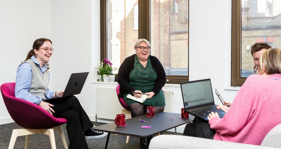 Several members of staff sitting around a communal working area in the Blood Cancer UK office, smiling and holding notebooks.
