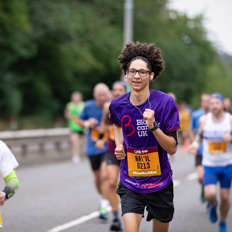 A woman running on a road wearing a purple blood cancer UK t shirt during a marathon.