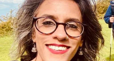 Profile photo of psychotherapist Surabhi, who is outside in the sun, wearing glasses and smiling.