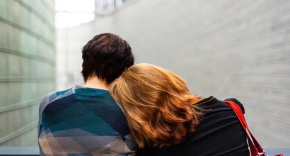 Two women shot from behind in a hospital setting: a woman with red hair lays her head on a dark-haired woman's shoulder