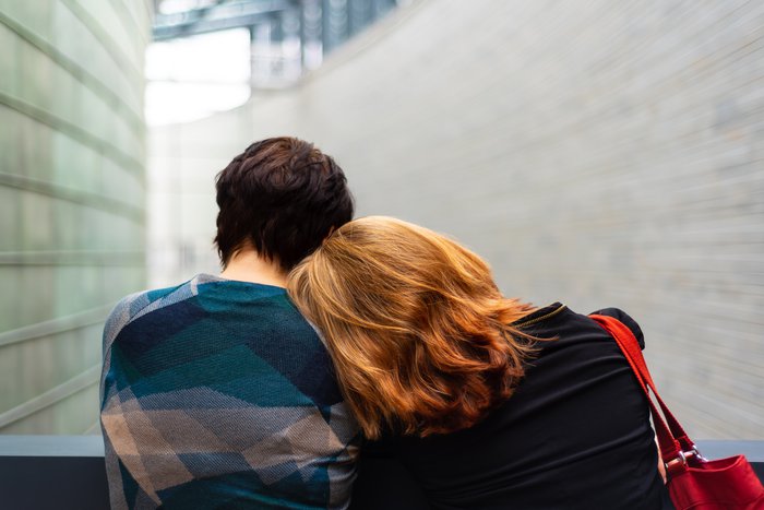 Two women shot from behind in a hospital setting: a woman with red hair lays her head on a dark-haired woman's shoulder
