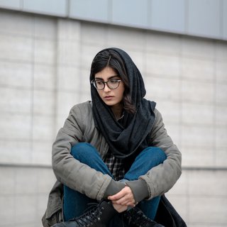A young woman wearing a headscarf sits on a wall looking sad and thoughtful.
