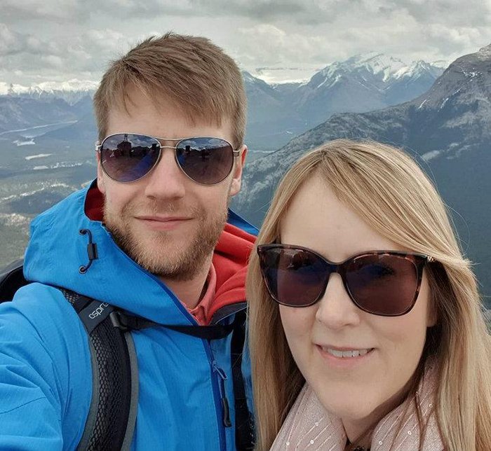 A young couple pose for a phone selfie on holiday, with snow-capped mountains in the background.