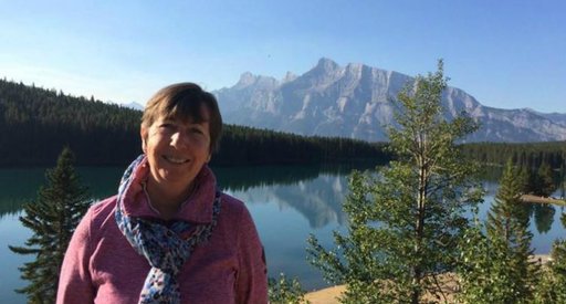 A woman poses for a holiday snap in front of a lake scene on a sunny day, with mountains in the background.