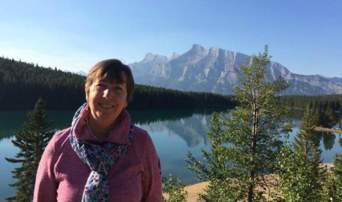 A woman poses for a holiday snap in front of a lake scene on a sunny day, with mountains in the background.
