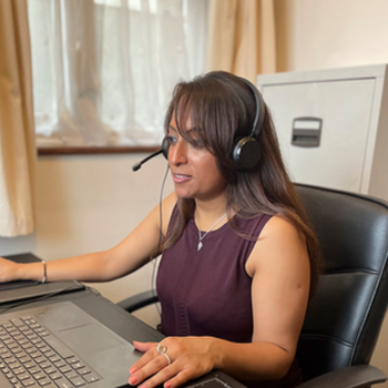 A woman takes a call on a headset as she works from home, looking at the computer in front of her. She's seated at a leather office chair with a filing cabinet and window behind her.